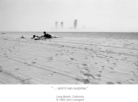People lay out on the sand Long Beach while fog covers most of the cityscape in front of them.