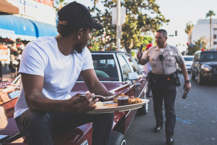 Kendrick Lamar eating outside of Roscoe's as a cop inspects a car and write's him a ticket in Los Angeles. (Photo by Jeff Lombardo)