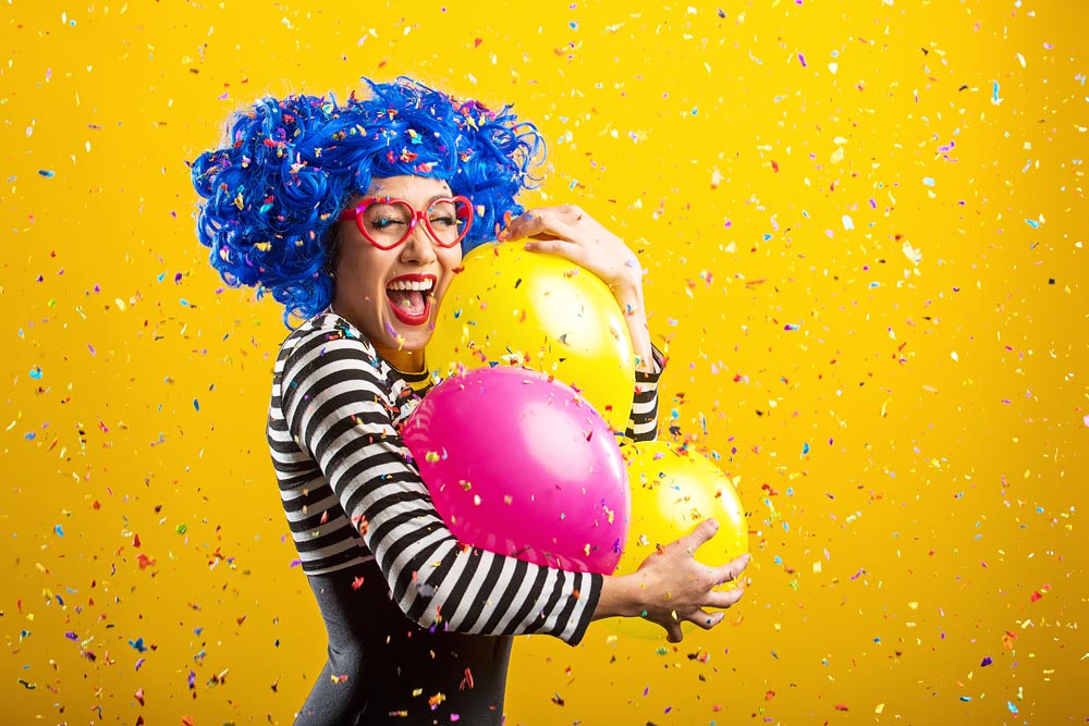 Woman with blue hair and red glasses wearing black and white striped shirt, standing in front of yellow background, holding pink and yellow balloons with confetti falling around her